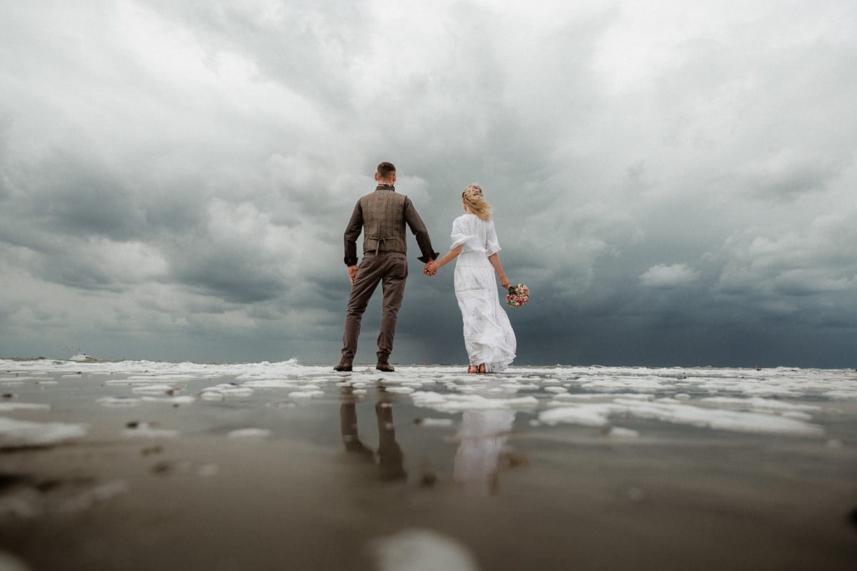 Handhaltendes Hochzeitspaar steht im Watt vor dramatischen Wolken auf Norderney.