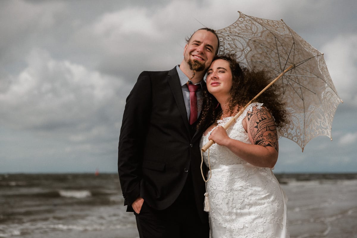Braut im Boho Look und Schirm mit ihrem Ehemann vor der Nordsee am Inselstrand.