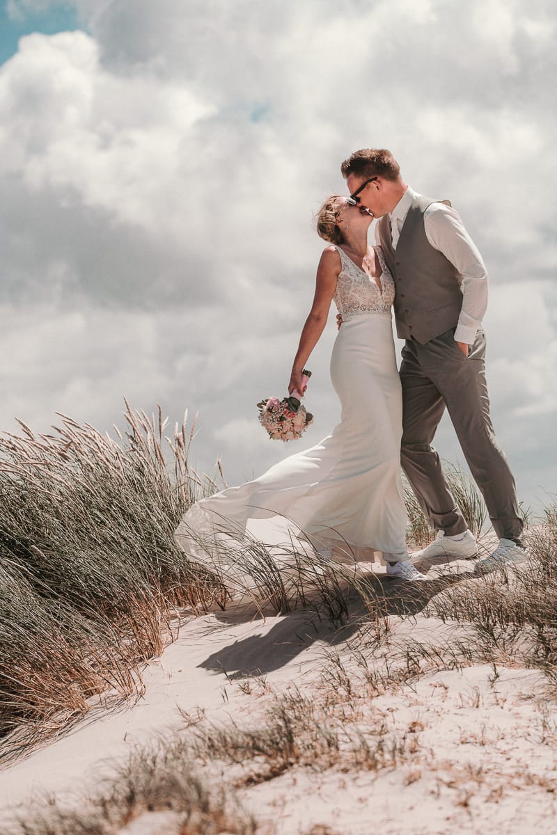 Brautkleid im Mermaid Look in den Duenen der Nordseeinsel Norderney mit ihrem Ehemann.