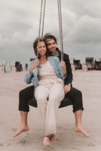 Junges Hochzeitspaar in legerem Look schaukelt zusammen am Strand vor den Strandkoerben.