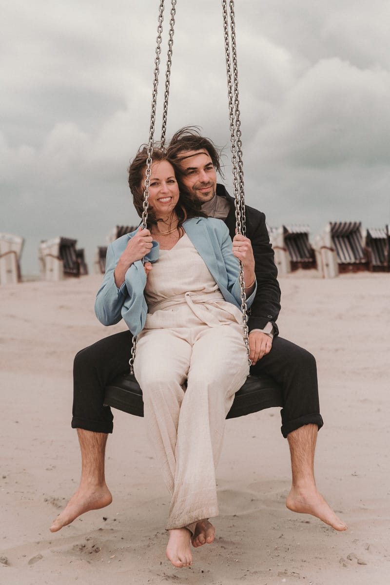 Junges Hochzeitspaar in legerem Look schaukelt zusammen am Strand vor den Strandkoerben.