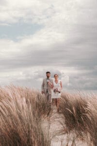 Eine Braut in kurzem Kleid spaziert mit ihrem Mann auf einem kleine Duenenweg auf der Nordseeinsel Norderney.