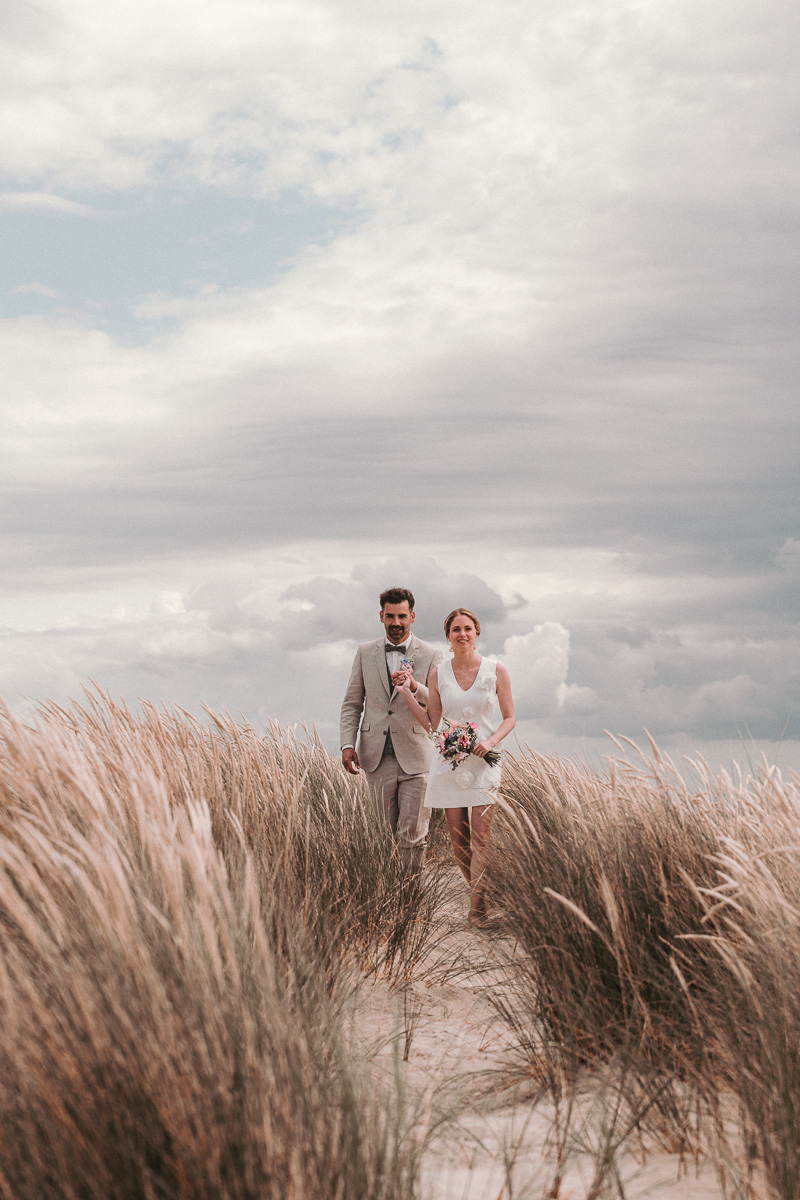 Eine Braut in kurzem Kleid spaziert mit ihrem Mann auf einem kleine Duenenweg auf der Nordseeinsel Norderney.