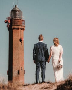 Der Leuchtturm von Norderney im Vordergrund ist ein Hochzeitspaar handhaltend.
