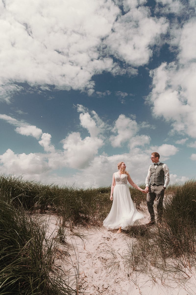 unter einem wunderschoenen Inselhimmel spaziert ein Brautpaar durch die Duenen an der Nordsee in Ostfriesland.