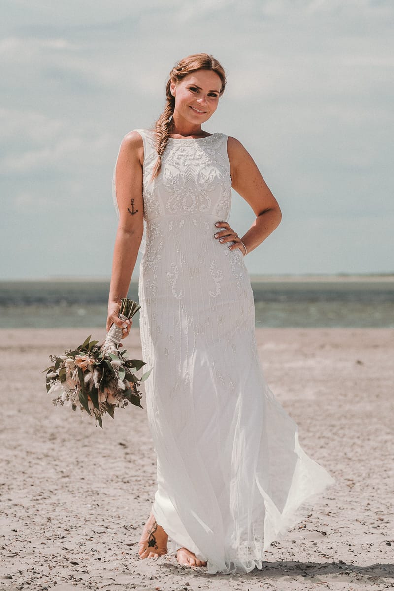 Vor der Wasserkante stehende Braut mit langem Hochzeitskleid und Brautstrauss.