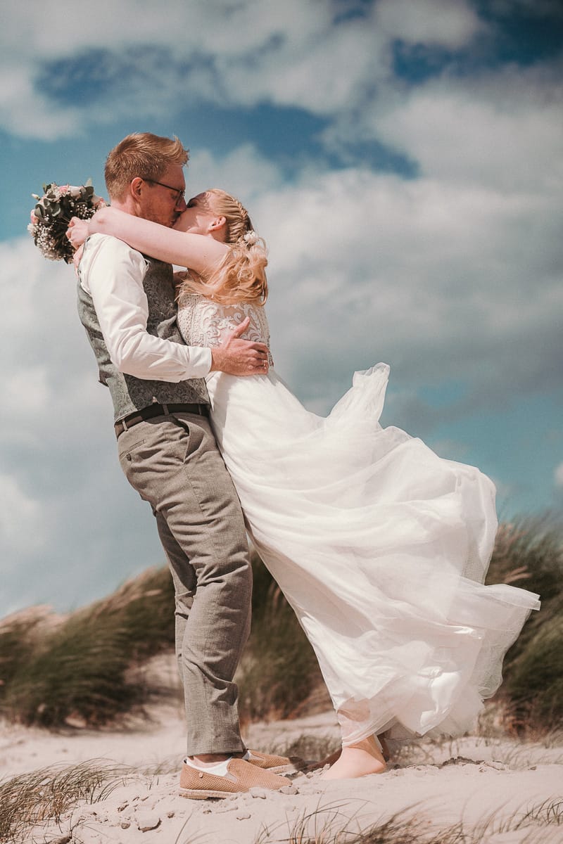 Die Braut kuesst den Braeutigam an der struermischen Nordsee mit wehendem Kleid.