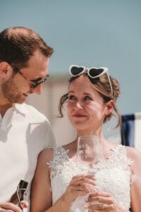 Braut mit Herzchenbrille vor dem Badekarren Norderney mit Sektglas schaut zu Ihrem Brautigam.